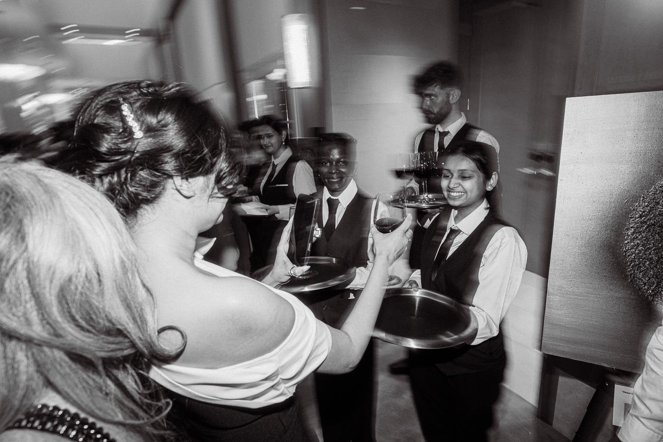Black and white image of a server handing a glass of wine to a woman at an event, captured with a motion blur effect, showing a lively and bustling atmosphere.