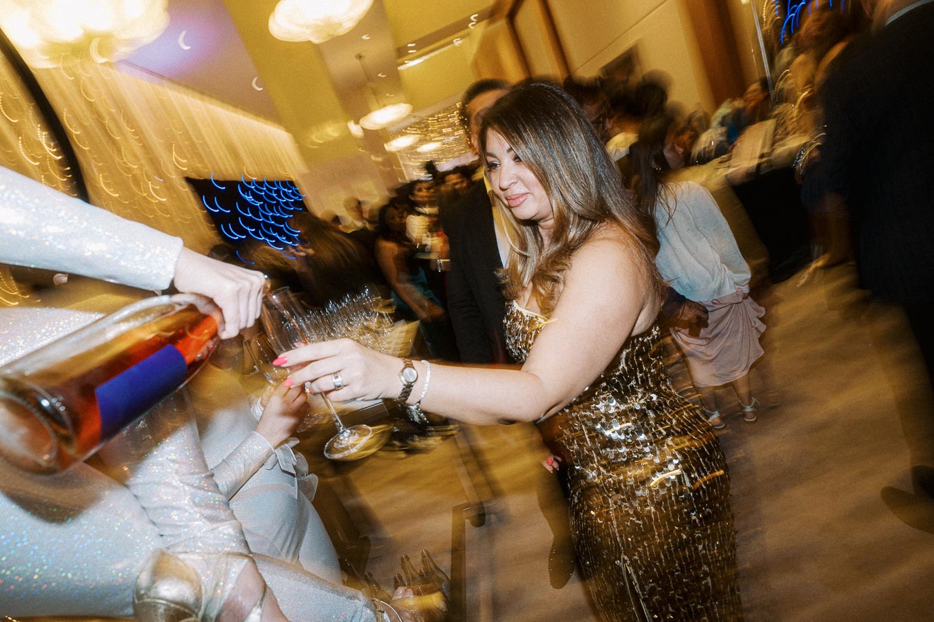 A woman in a sequined dress receives champagne at a lively event, captured in vibrant motion, with people socializing in a warmly lit venue.