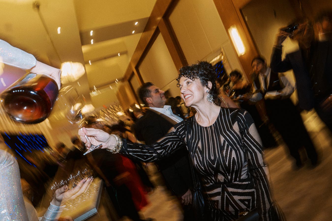 Woman in elegant black dress cheerfully receiving wine pour at a lively event, with blurred background of other guests and warm lighting.