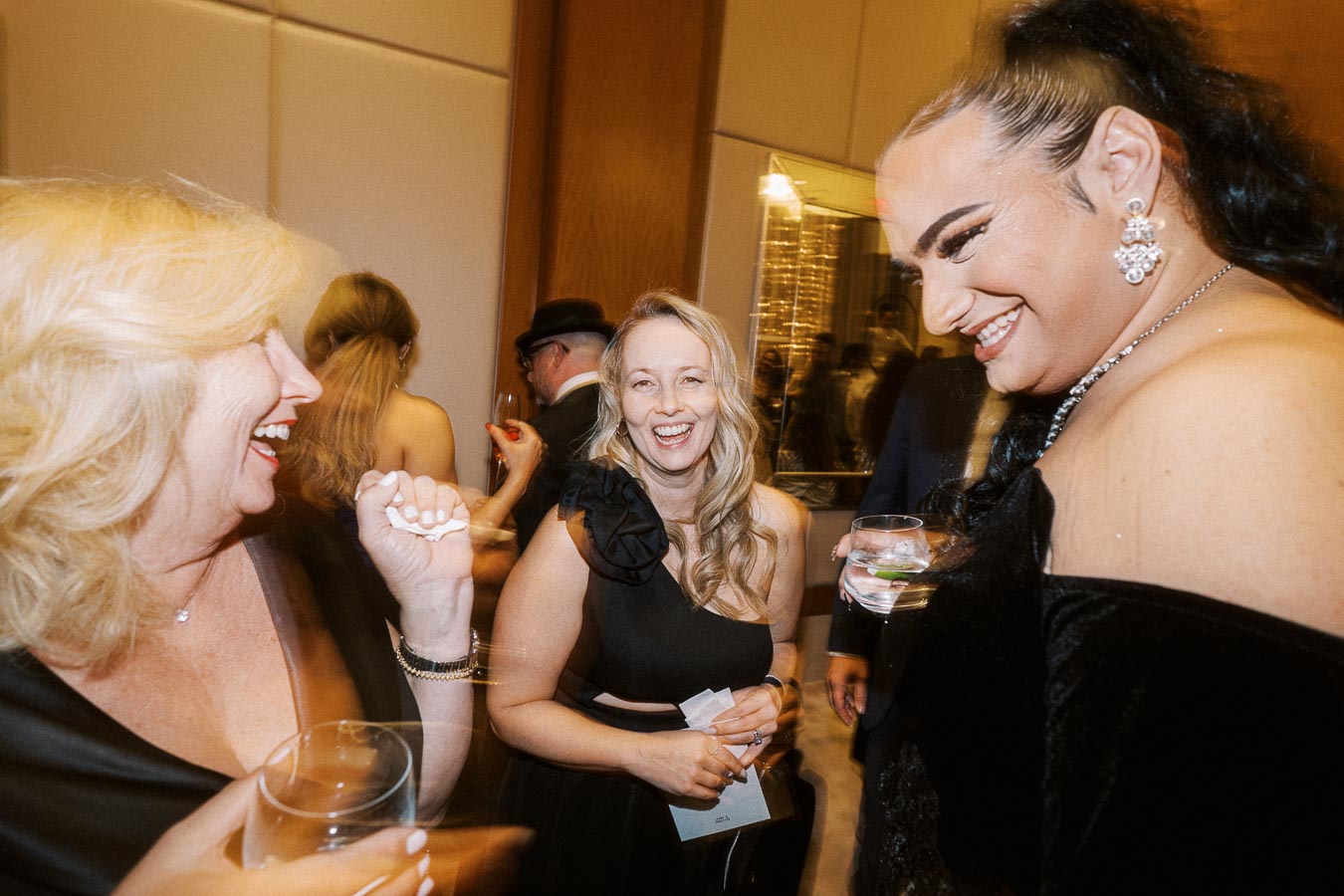 A group of elegantly dressed individuals laughing and enjoying a social gathering, each holding a drink, in a warmly lit event space.