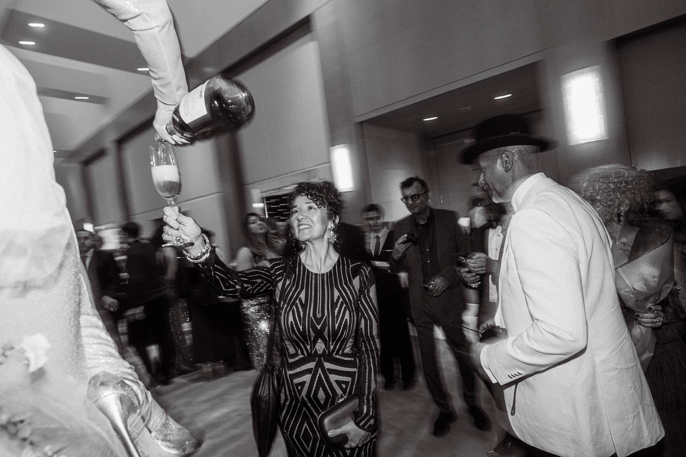 A woman in an elegant dress holds up a glass of champagne being poured from a large bottle at a lively party, surrounded by guests in formal attire, exuding a joyful and celebratory atmosphere.