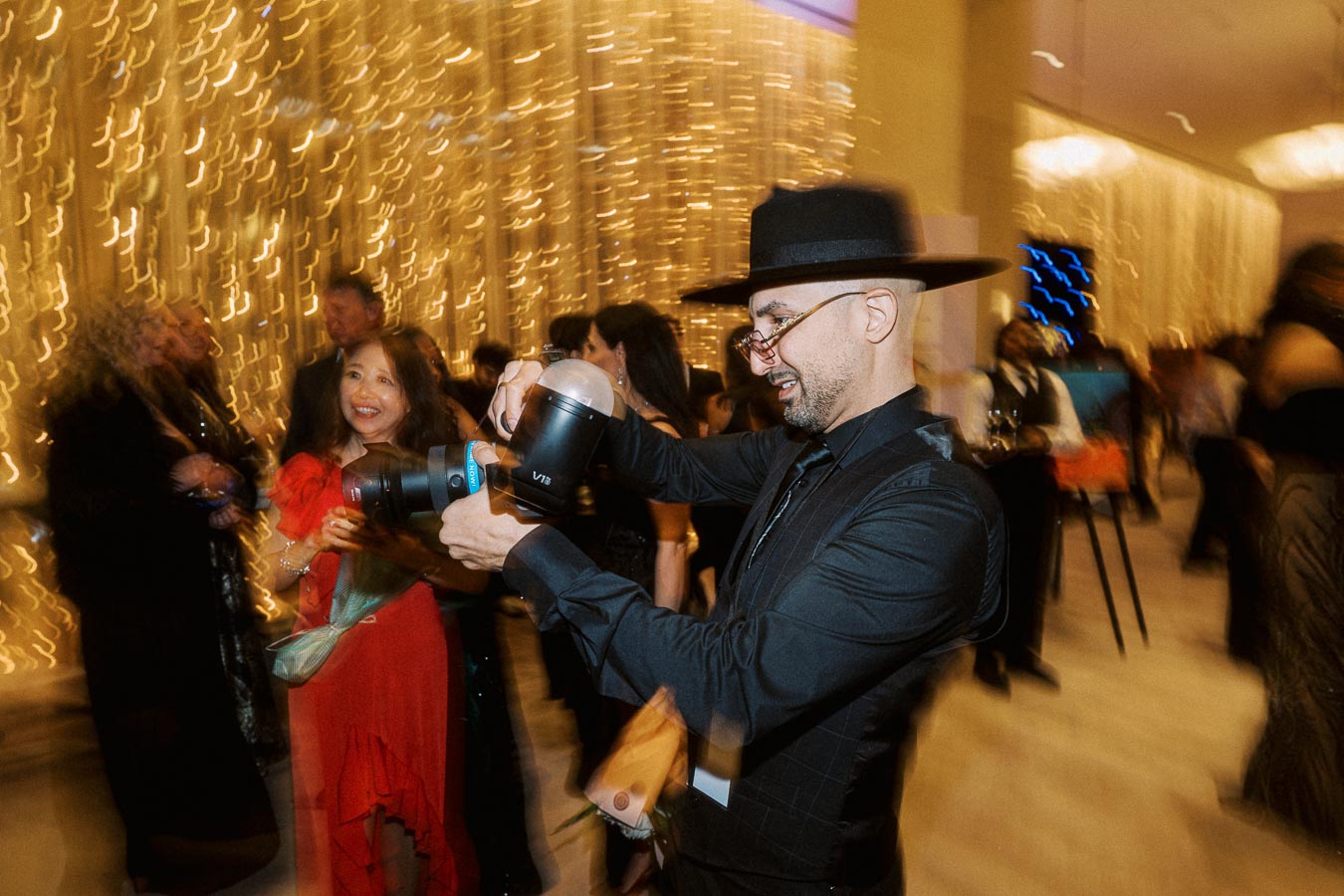 Man wearing black hat taking photos at a lively event with blurred lights and guests in the background.