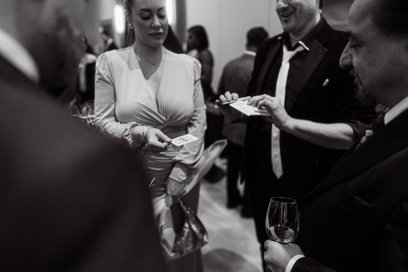 A black and white photo capturing a lively scene at a formal event, where attendees in elegant attire engage in a card game, as one person skillfully demonstrates a card trick to others gathered around, with wine glasses in hand.