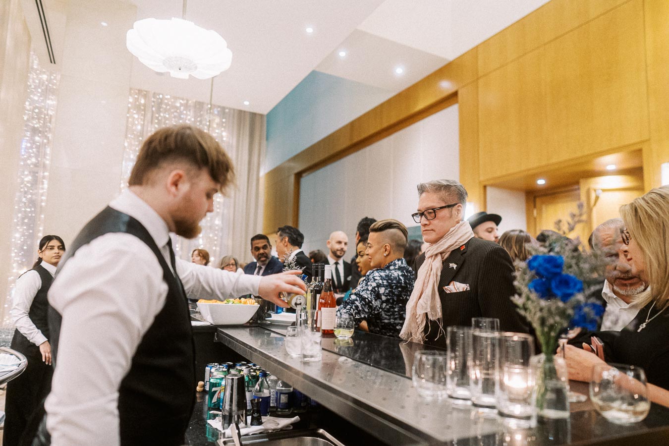 A bartender serves drinks to well-dressed guests at a stylish indoor event. The bar is adorned with flowers and lit with soft lighting, creating an elegant and sophisticated atmosphere.