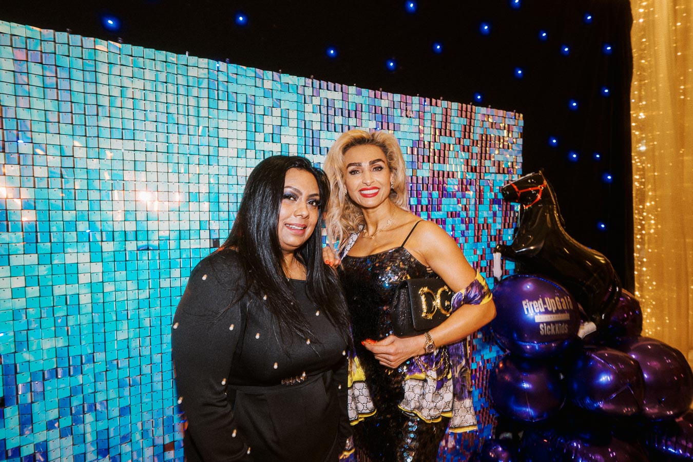 Two women at a glamorous event, wearing elegant outfits, stand in front of a shimmering blue sequin backdrop adorned with blue lights. They are smiling for the camera, accompanied by decorative purple balloons to their side.