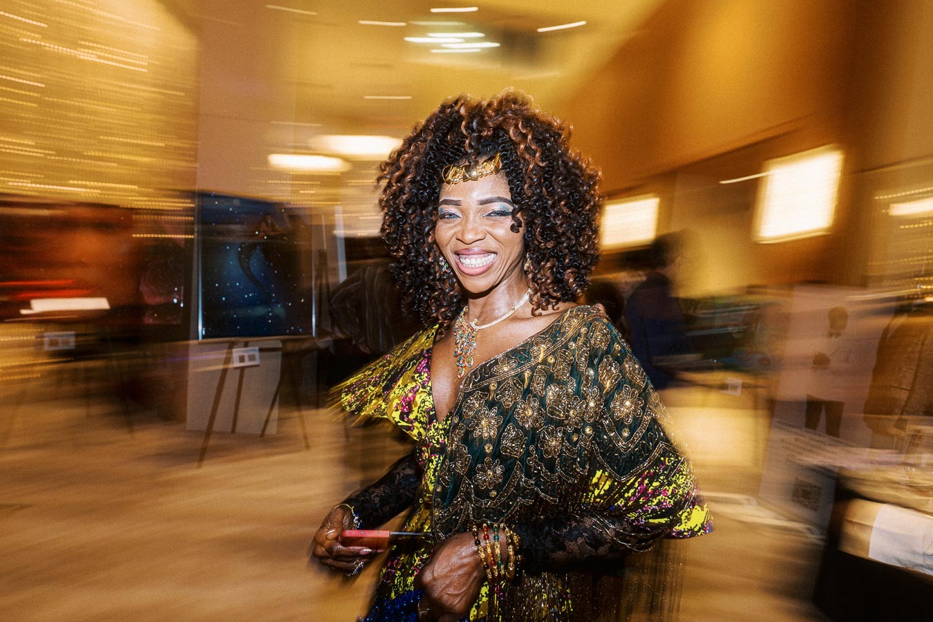 Smiling woman in elegant, colorful attire with ornate patterns, holding a phone, surrounded by a blurred, dynamic event setting.