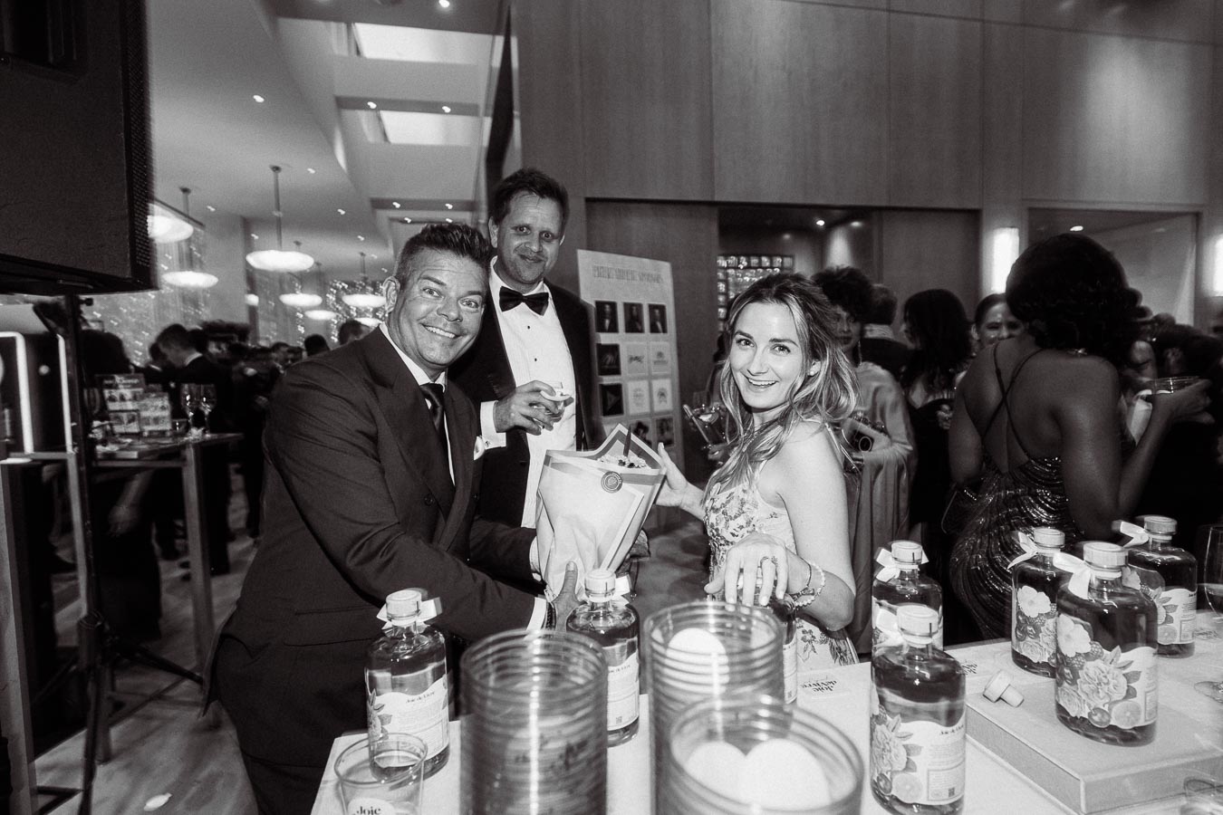 Black and white photo of a lively event with elegantly dressed attendees enjoying drinks at a well-stocked bar, featuring a man and woman smiling directly at the camera amidst a bustling, upscale atmosphere.
