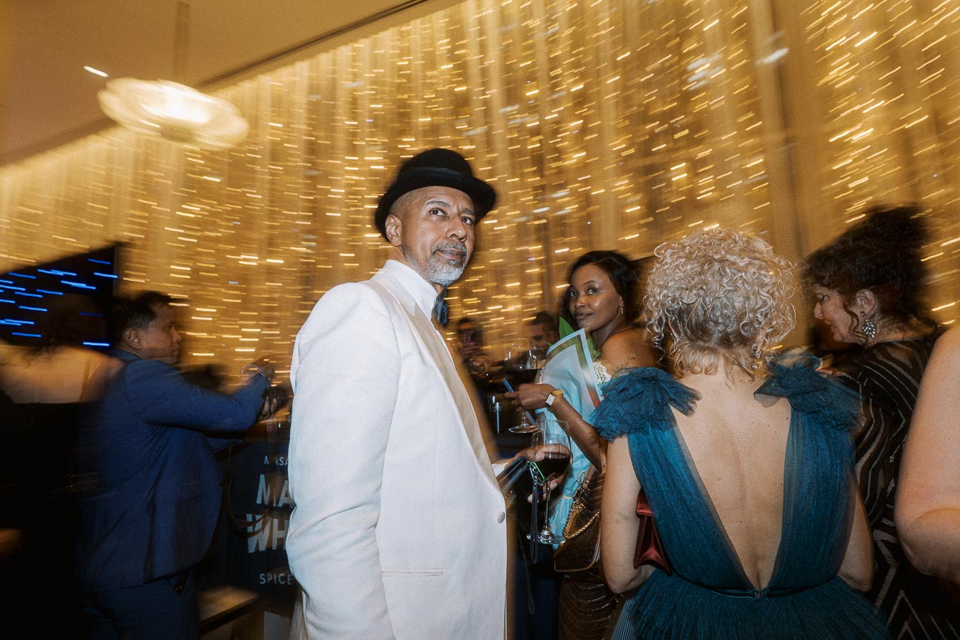 A lively evening gala with elegantly dressed attendees enjoying a sophisticated ambiance, featuring a well-dressed man in a white suit and black hat amidst a crowded room with golden light accents.