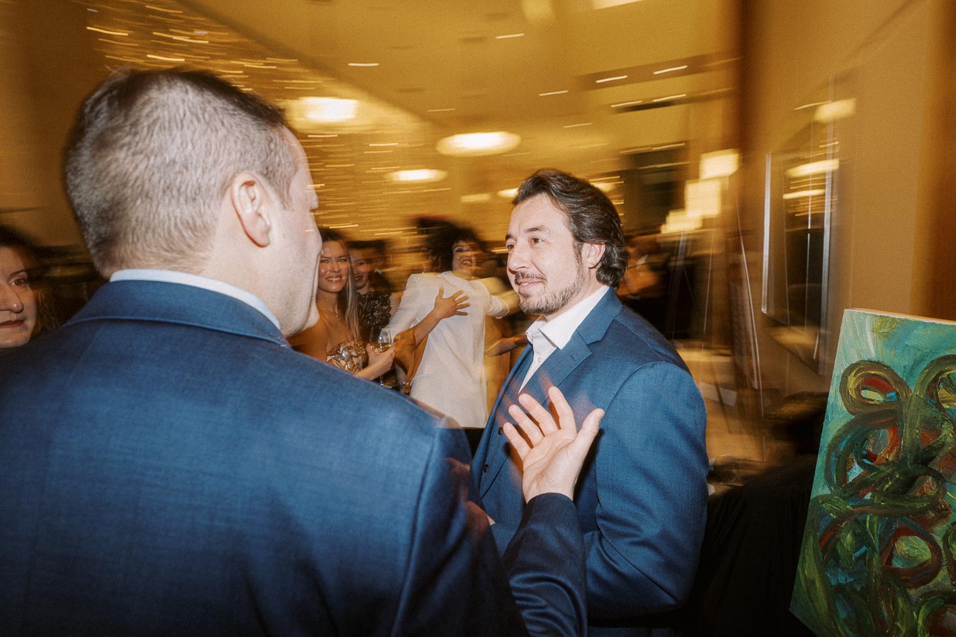 A group of people in formal attire socializing at an elegant indoor event, with blurred lights in the background creating a lively atmosphere. A colorful painting is visible in the foreground.