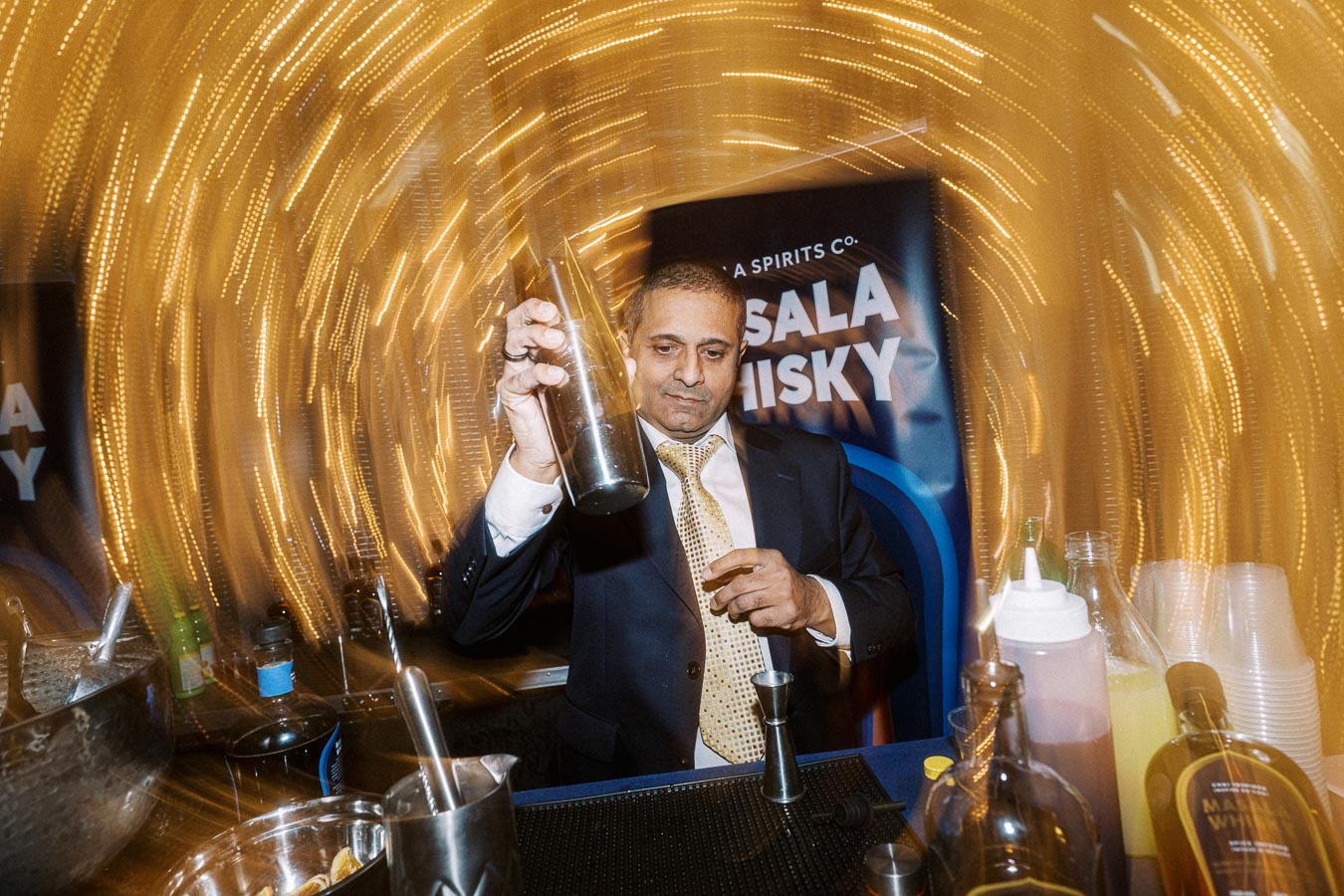 Bartender expertly mixing a cocktail with a shaker at a vibrant event, surrounded by bottles and a backdrop promoting masala whisky. Bright motion lights create a dynamic atmosphere.