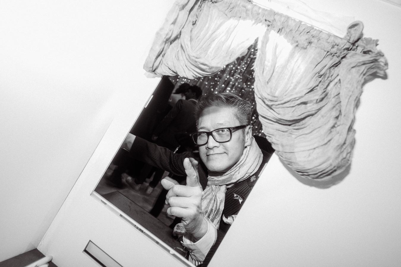 A person wearing glasses and a scarf smiling and pointing through a decorated window, captured in black and white with a festive background.
