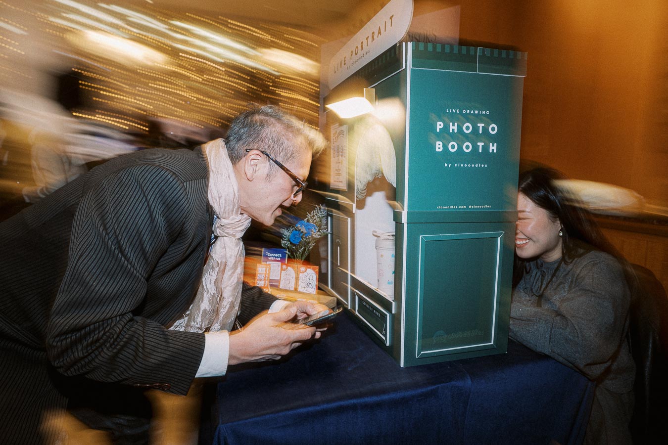 Man interacting with a live drawing photo booth at an event, holding his phone, while a woman artist inside smiles, capturing the scene in a dynamic, blurred environment.