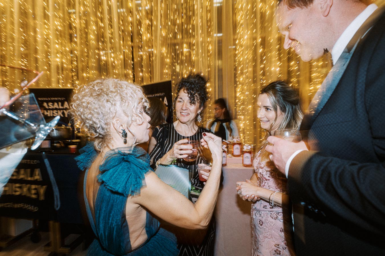 A lively social gathering with elegantly dressed guests enjoying drinks and conversation under warm string lights.