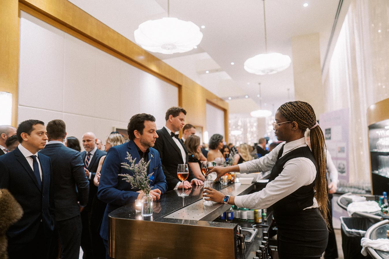 Elegant hotel event bar scene with a diverse crowd of formally dressed people socializing; bartender serving drinks, ambient lighting in stylish venue.