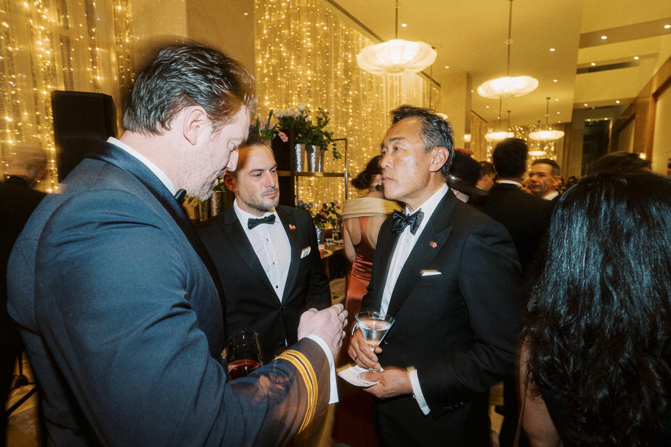 Elegant party setting with guests in formal attire engaging in conversation, surrounded by ambient golden lights and lavish decor.