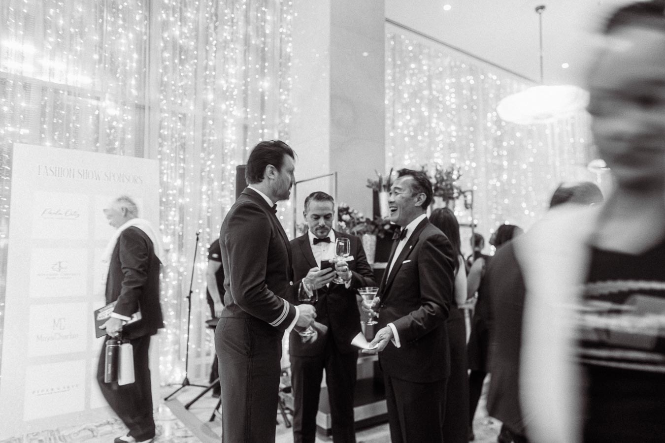 Black and white image of a formal gathering, featuring elegantly dressed individuals in conversation, holding drinks at a sophisticated event with a backdrop of twinkling lights and a display board.