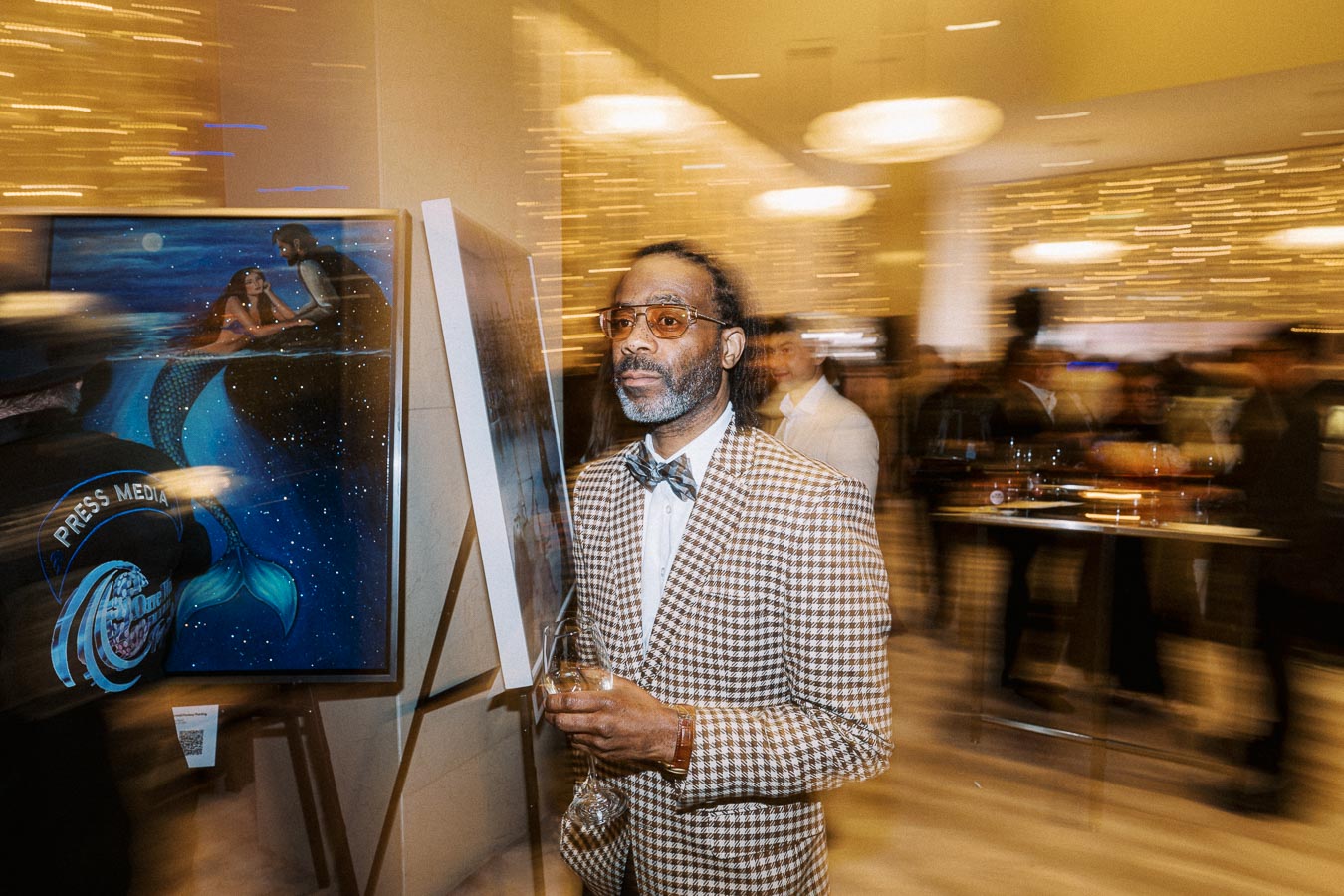 A man in a checkered suit holding a drink stands in an art gallery, surrounded by blurred motion, with a painting of a mermaid on an easel in the background.