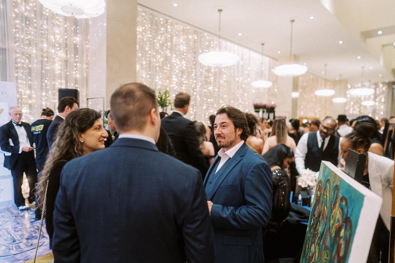 Elegant gala event with guests mingling in formal attire under sparkling lights, featuring artwork on display.