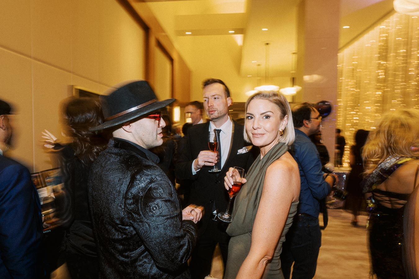 Group of elegantly dressed people socializing at a sophisticated indoor event, with one woman in focus holding a drink and smiling at the camera.