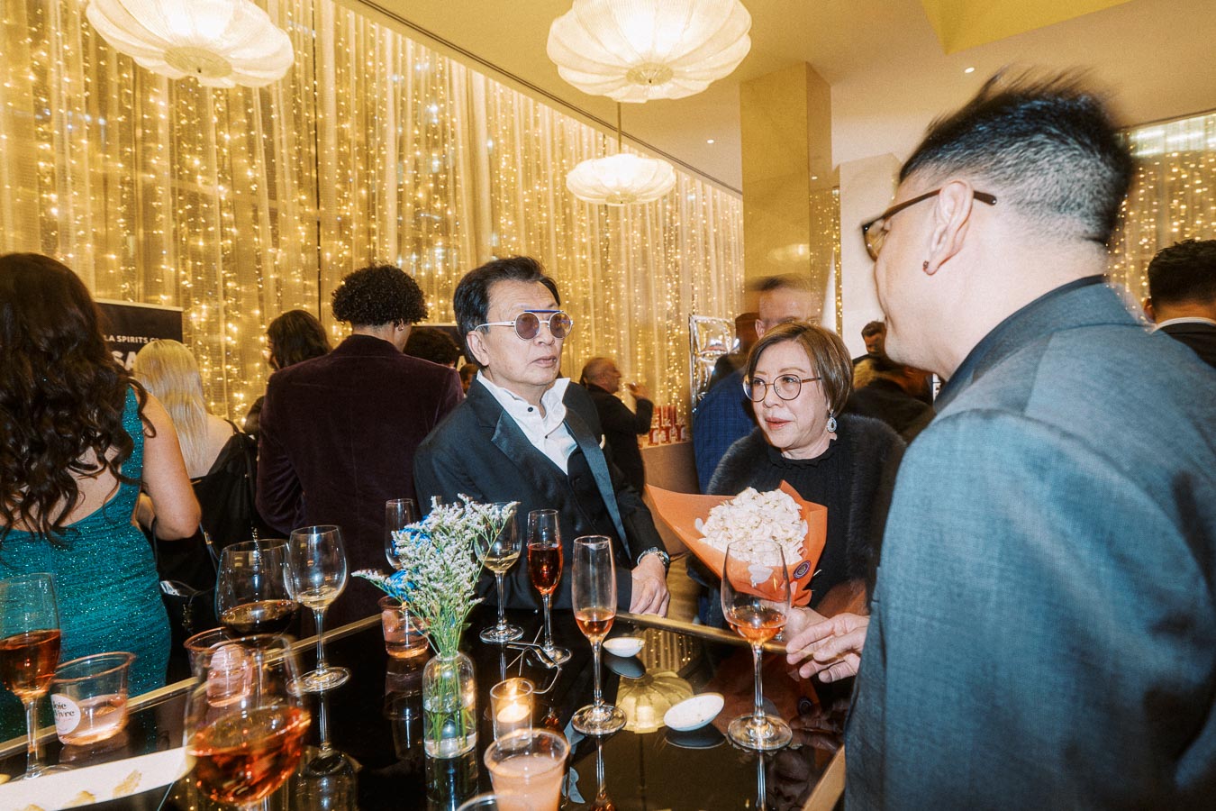 A lively cocktail party scene with elegantly dressed attendees mingling near a decorated table with drinks and floral arrangements, amidst a backdrop of warm fairy lights.