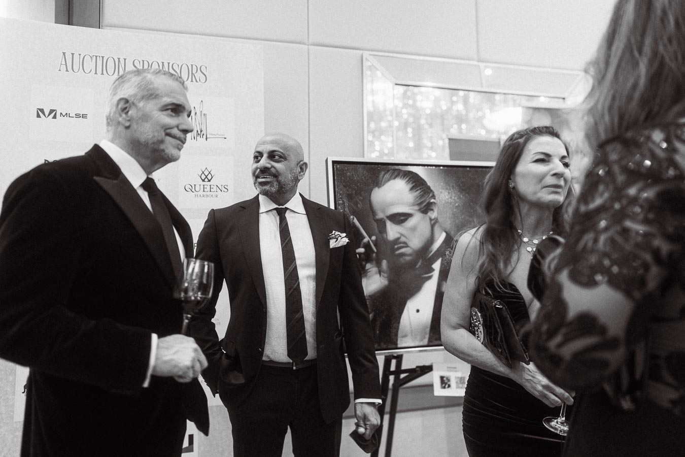 Black and white photo of elegantly dressed people at a formal event, with a portrait of a classic film icon in the background and auction sponsor details displayed, capturing the sophisticated atmosphere of an art gala.