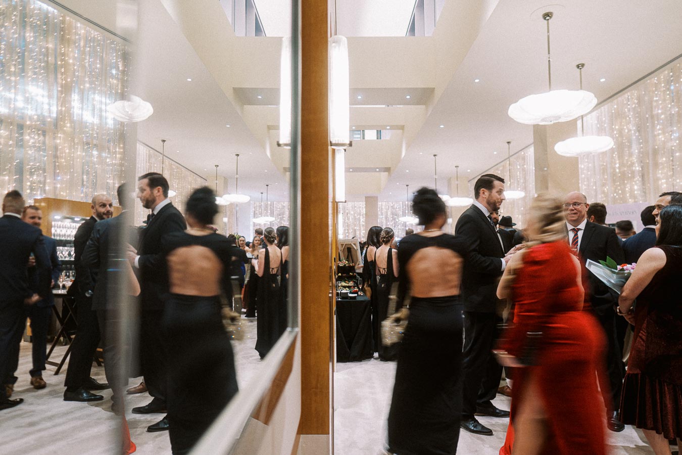 Elegant evening event with guests dressed in formal attire, mingling and socializing in a beautifully decorated venue with shimmering lights and modern architecture.