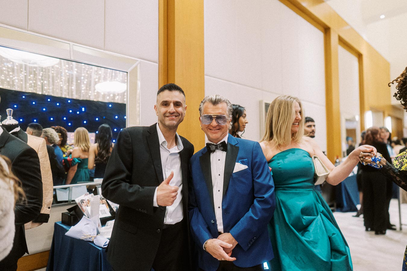 Two men dressed in formal suits, one giving a thumbs up, at an elegant event with people socializing in the background.