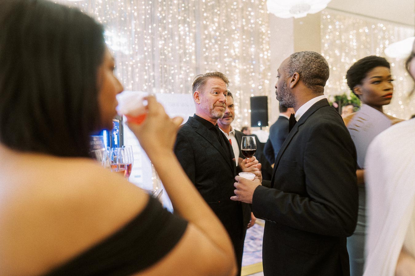 Guests at a formal event enjoying drinks and conversation in a beautifully lit venue with sparkling lights in the background.
