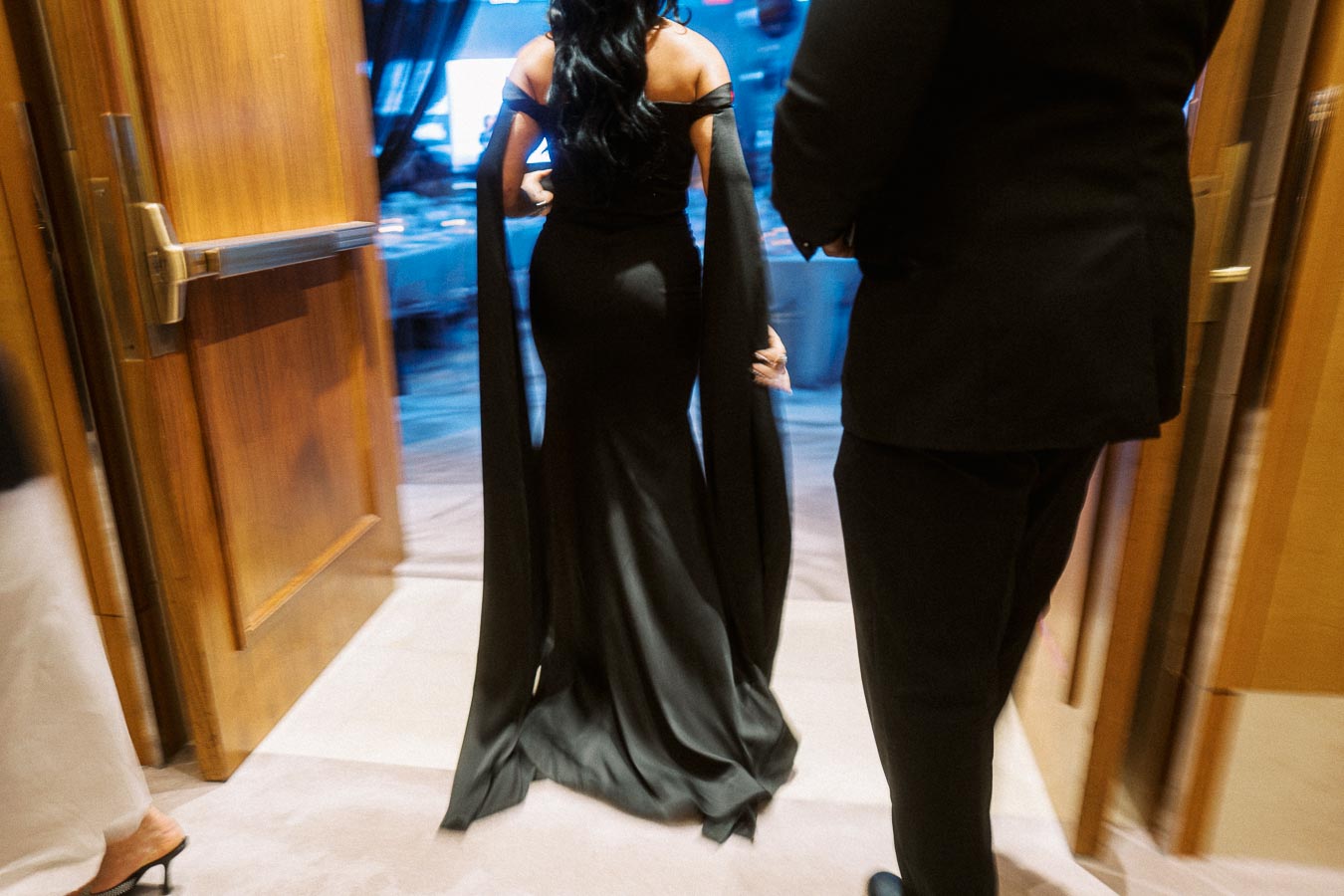 Elegant woman in a long black gown entering a formal event through wooden double doors, accompanied by someone in a dark suit, creating an atmosphere of sophistication and style.
