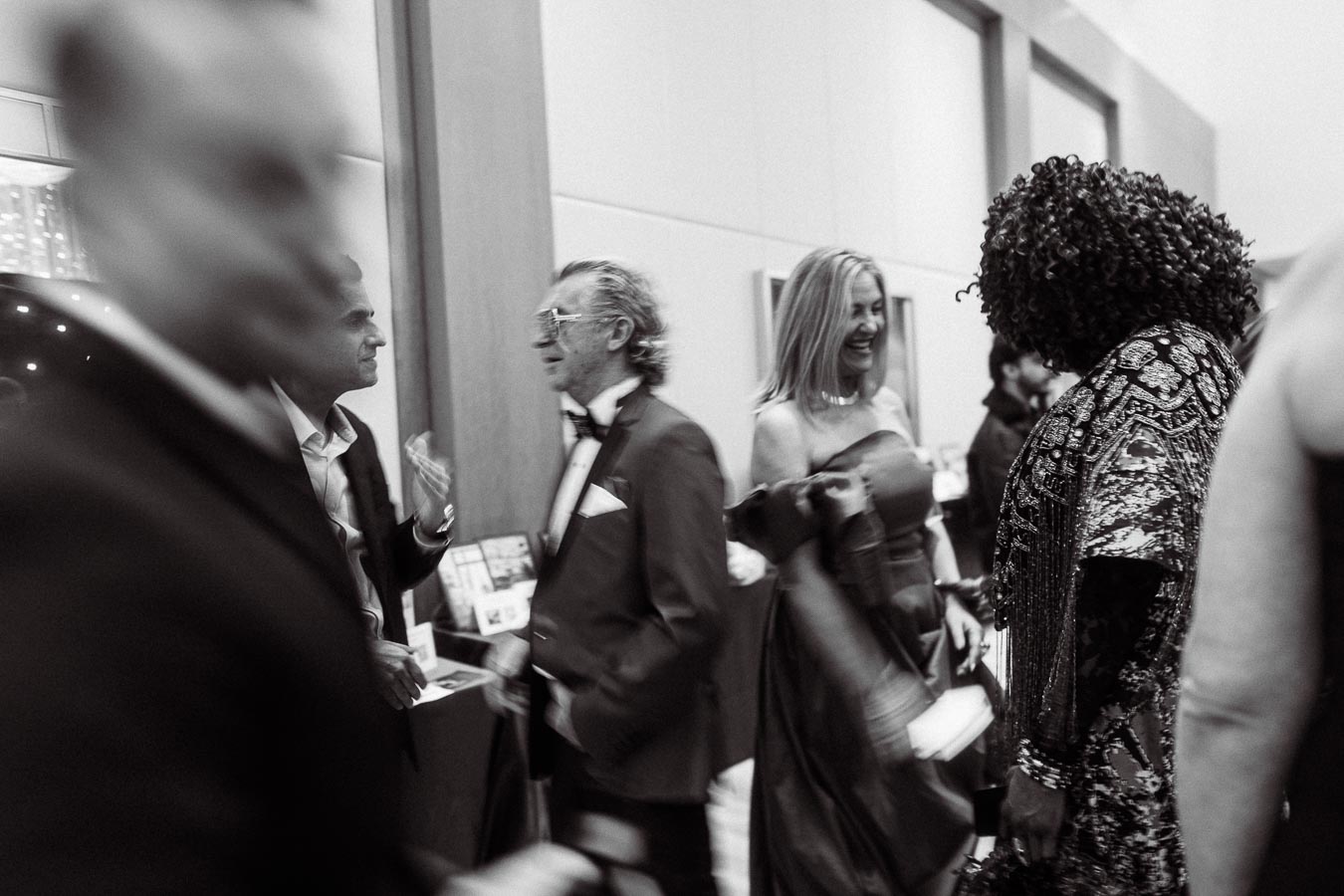 Black and white image of an elegant gathering, featuring people in formal attire at a sophisticated event, engaging in conversation and sharing laughter.