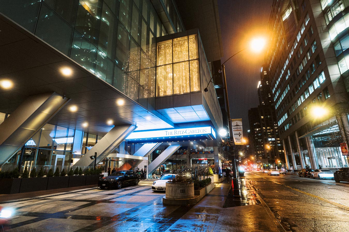 Nighttime view of a modern hotel entrance with bright lights, showcasing The Ritz-Carlton. Wet street reflects city lights, with cars parked and buildings flanking the scene, capturing a luxurious urban ambiance.