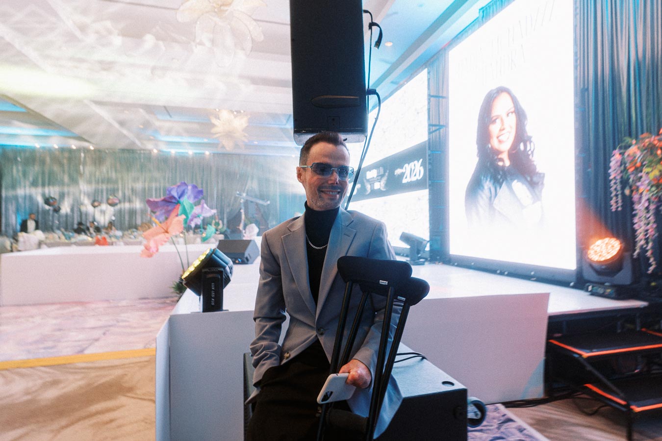 Man in a gray suit with crutches and sunglasses, smiling at an event with decorative flowers and a large screen in the background.
