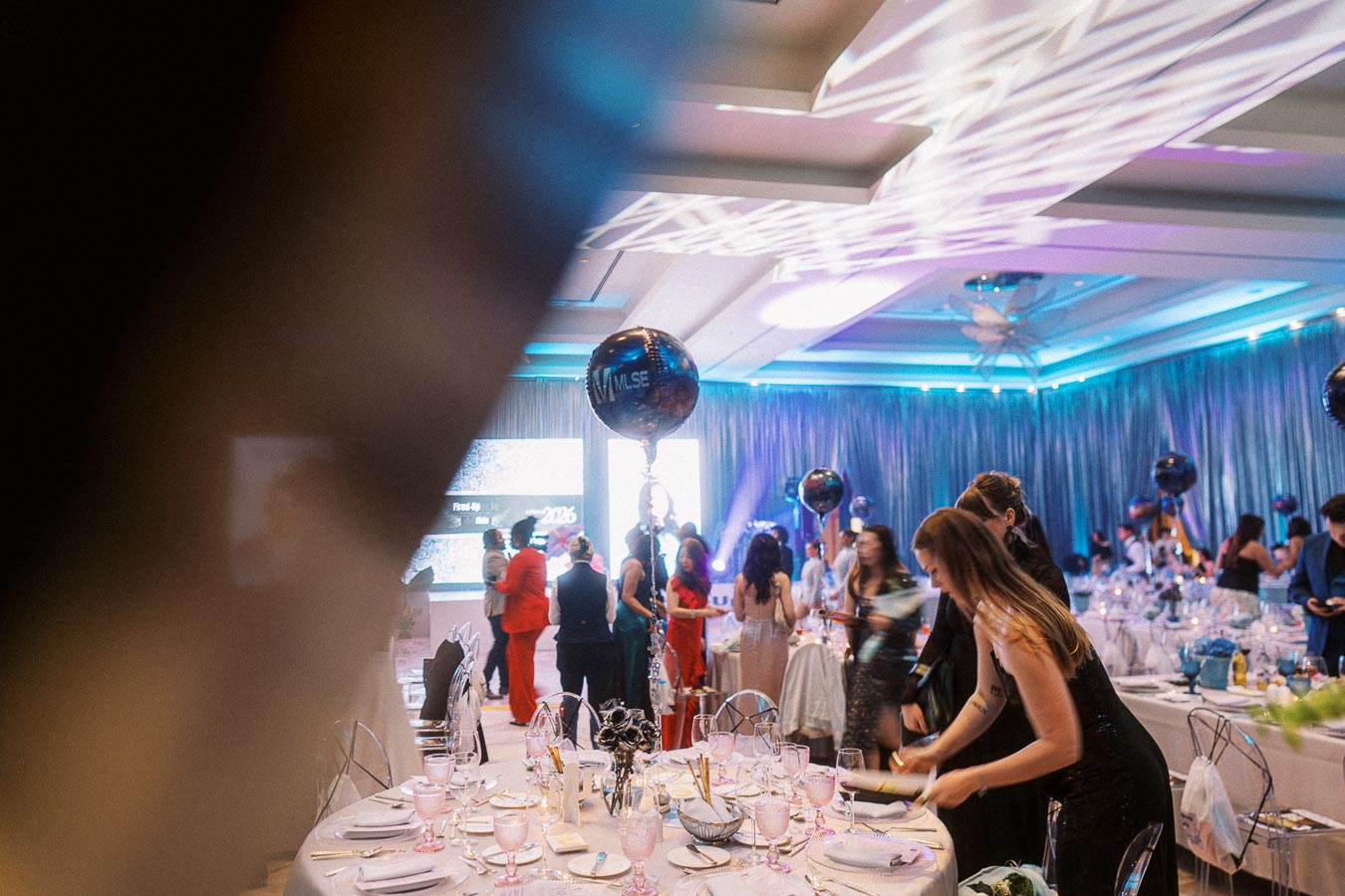 Elegant event hall with guests mingling near decorated tables, featuring blue lighting and festive balloons.