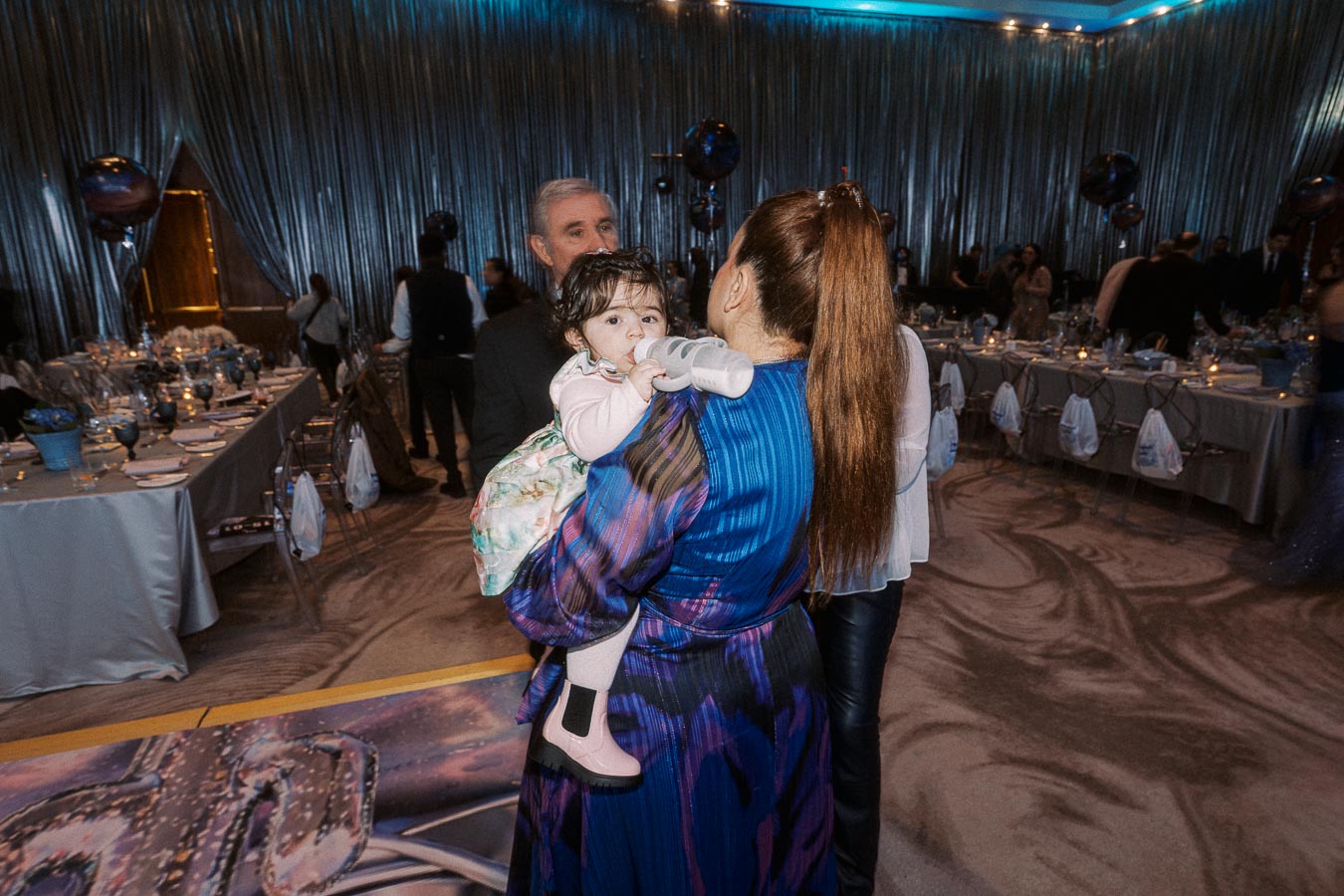A woman in a blue and purple dress is holding a baby drinking from a bottle at a formal event with elegantly set tables and guests in the background.