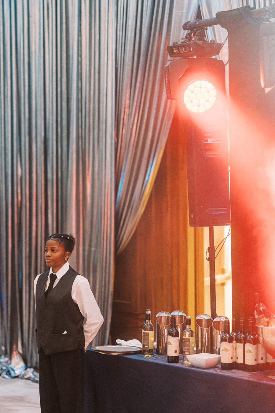 A formally dressed server stands beside a table set with wine bottles and glasses at an elegant event, with soft lighting and draped curtains in the background.