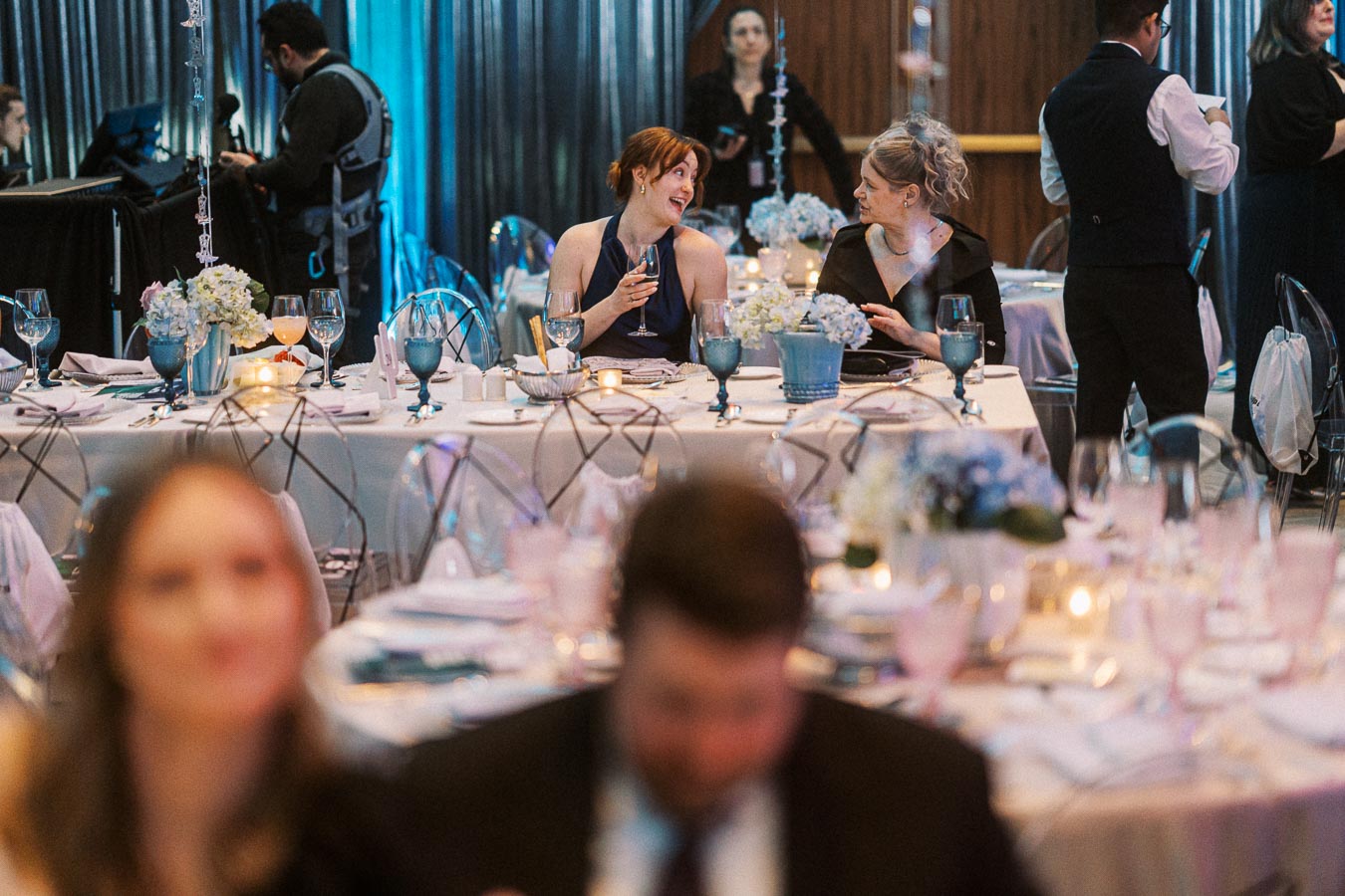 Elegant banquet hall setting with two women enjoying conversation at a beautifully decorated table, featuring floral centerpieces and soft lighting, while waitstaff and guests interact in the background.