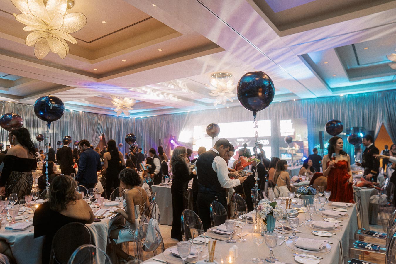 Elegant formal event in a luxurious ballroom, featuring guests in evening attire, decorated tables with fine dining settings, and ambient lighting with decorative balloons suspended from the ceiling.