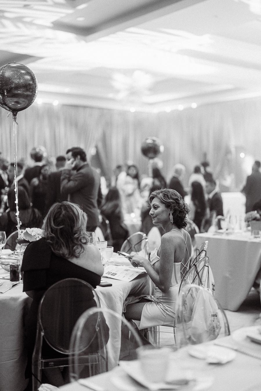 A black and white photo of a formal event with elegantly dressed women seated at a table. The bustling atmosphere includes guests mingling and decorative balloons, creating a sophisticated and lively setting.