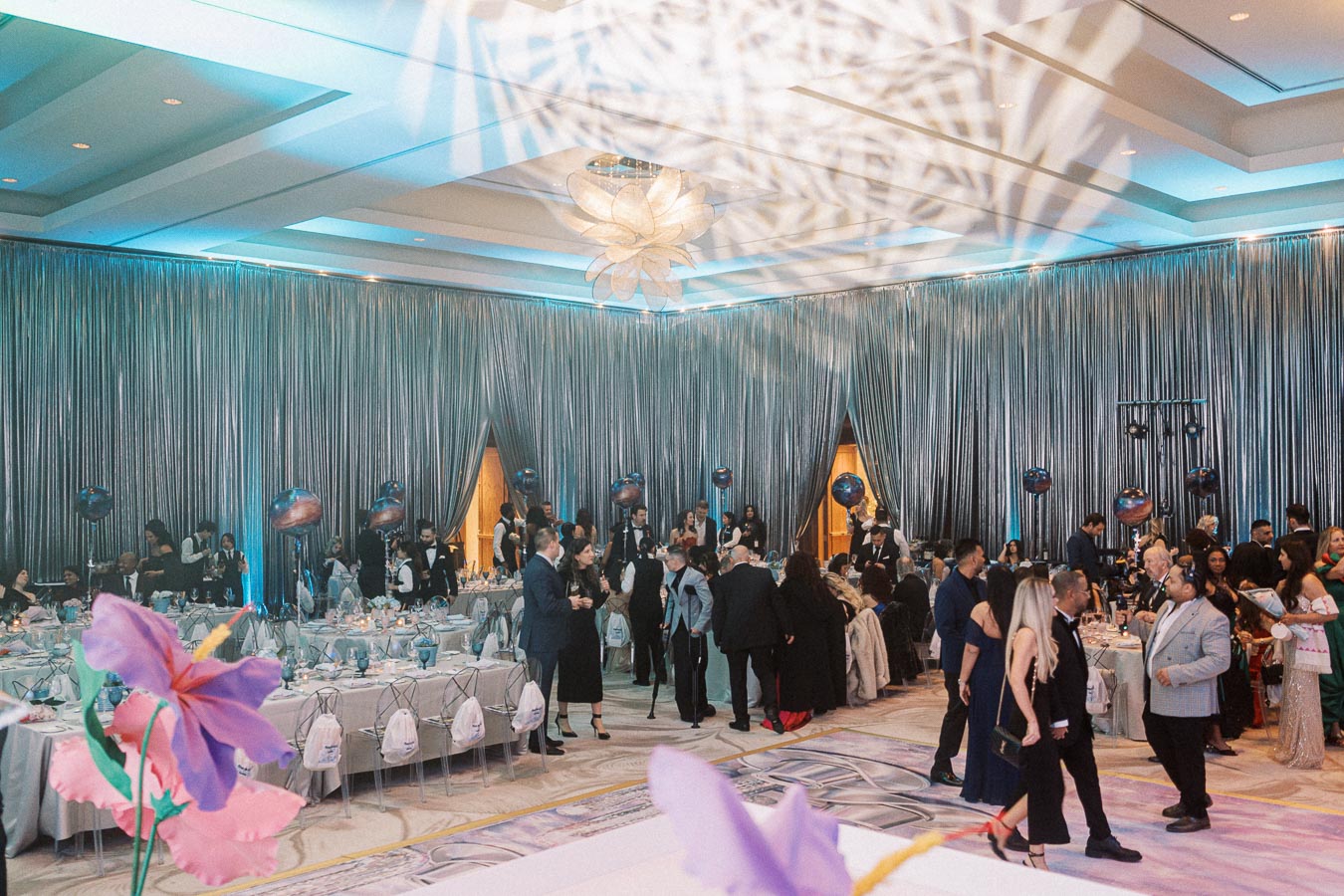 Luxurious indoor wedding reception with silver draped walls, elegantly set tables, and guests mingling in formal attire.