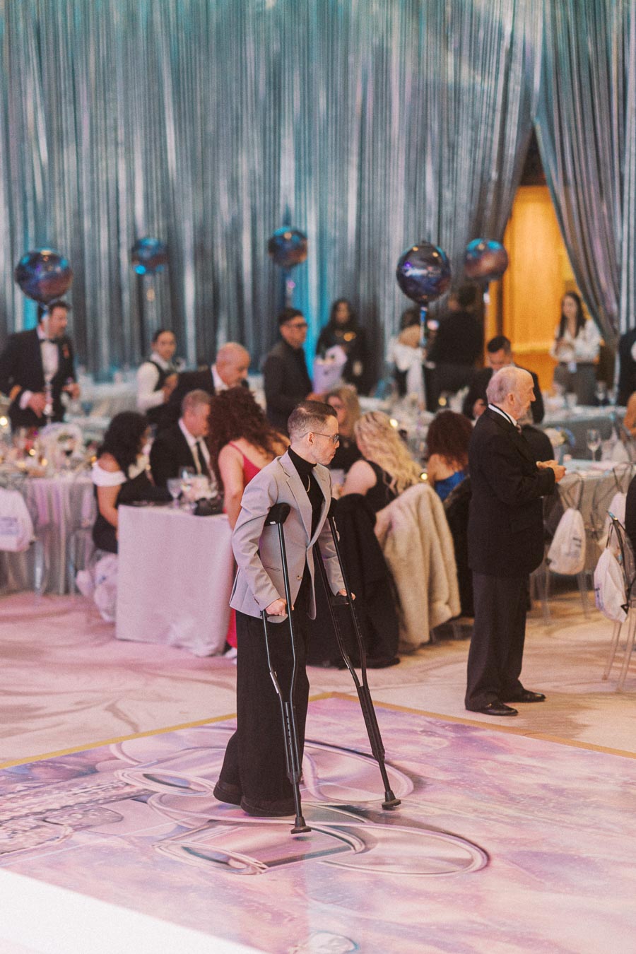 A person with crutches walking across a decorated dance floor at an elegant event, surrounded by guests in formal attire seated at tables with blue and silver decor.