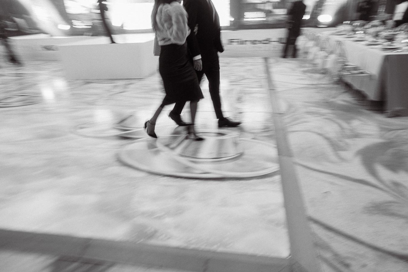 Blurred black and white image of a man and woman walking side by side on an elegant, patterned floor at a formal event.