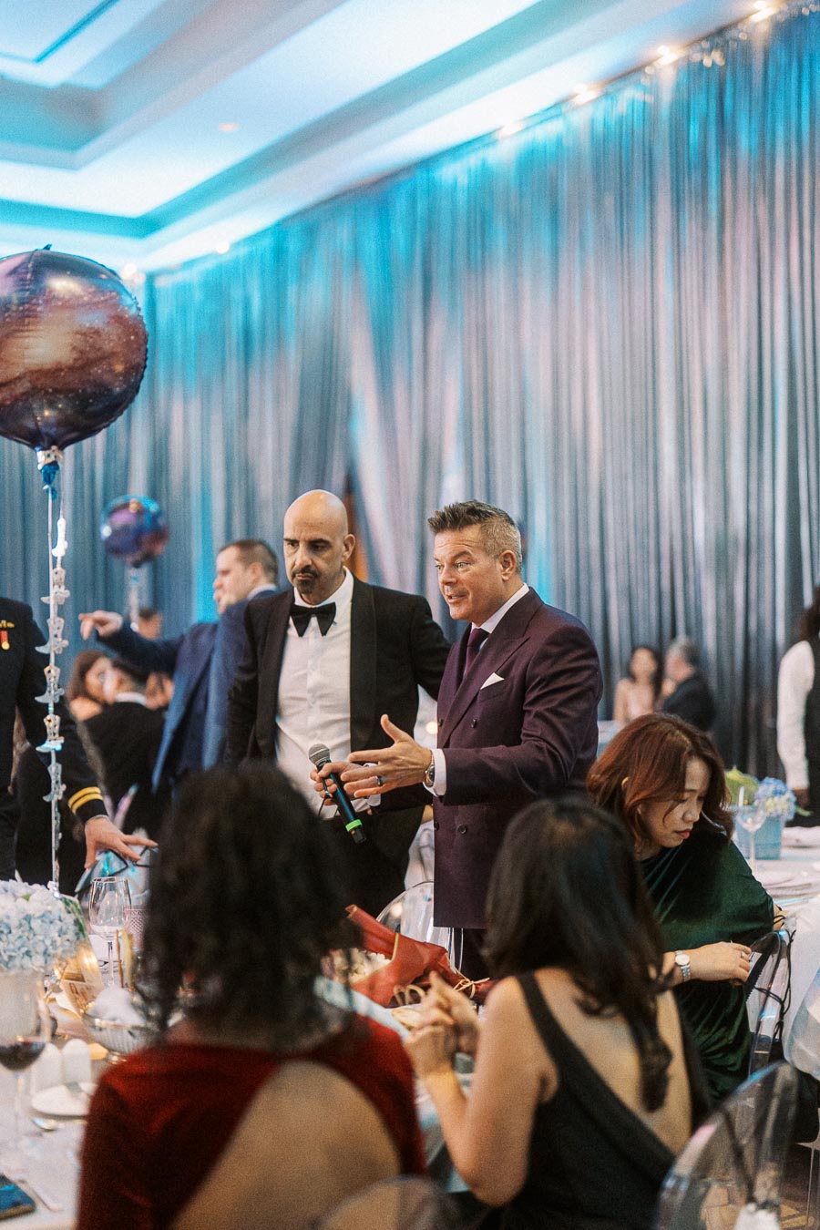Elegant dinner event with men in formal attire interacting with guests, microphone in hand, under soft blue lighting, decorative balloon centerpiece, and attendees seated at tables.