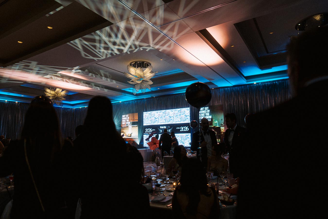 Elegant ballroom event with people in formal attire, under ambient lighting, featuring decorative floral ceiling lights and digital screens displaying dynamic content.