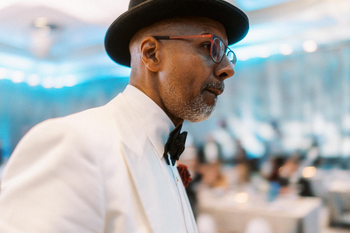A man in a stylish white suit, black bow tie, and hat at a formal event, with a blurred background of people and soft blue lighting.