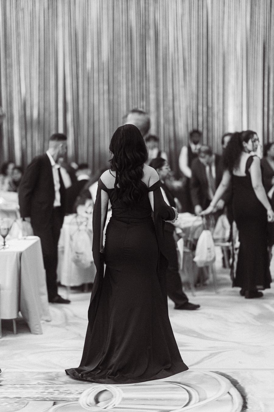 Black and white photo of a formal event, with an elegantly dressed woman in a long black gown walking through a bustling crowd of similarly attired guests, creating a sophisticated and lively atmosphere.