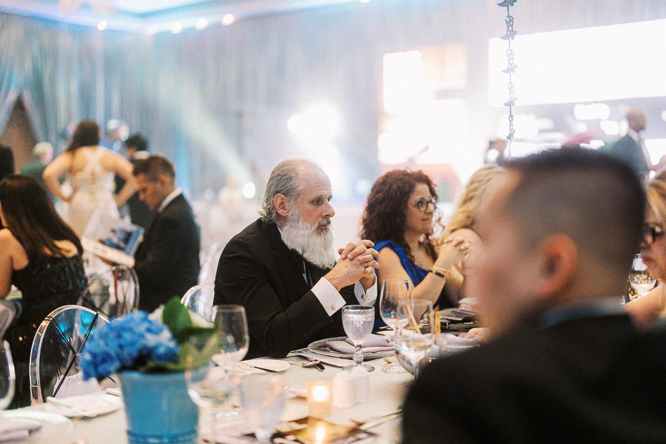 Elegant evening gala event with formally dressed guests seated at a beautifully decorated table, featuring a white-haired man with a beard in a suit, ambient lighting, floral arrangements, and glassware indicating a classy celebration atmosphere.