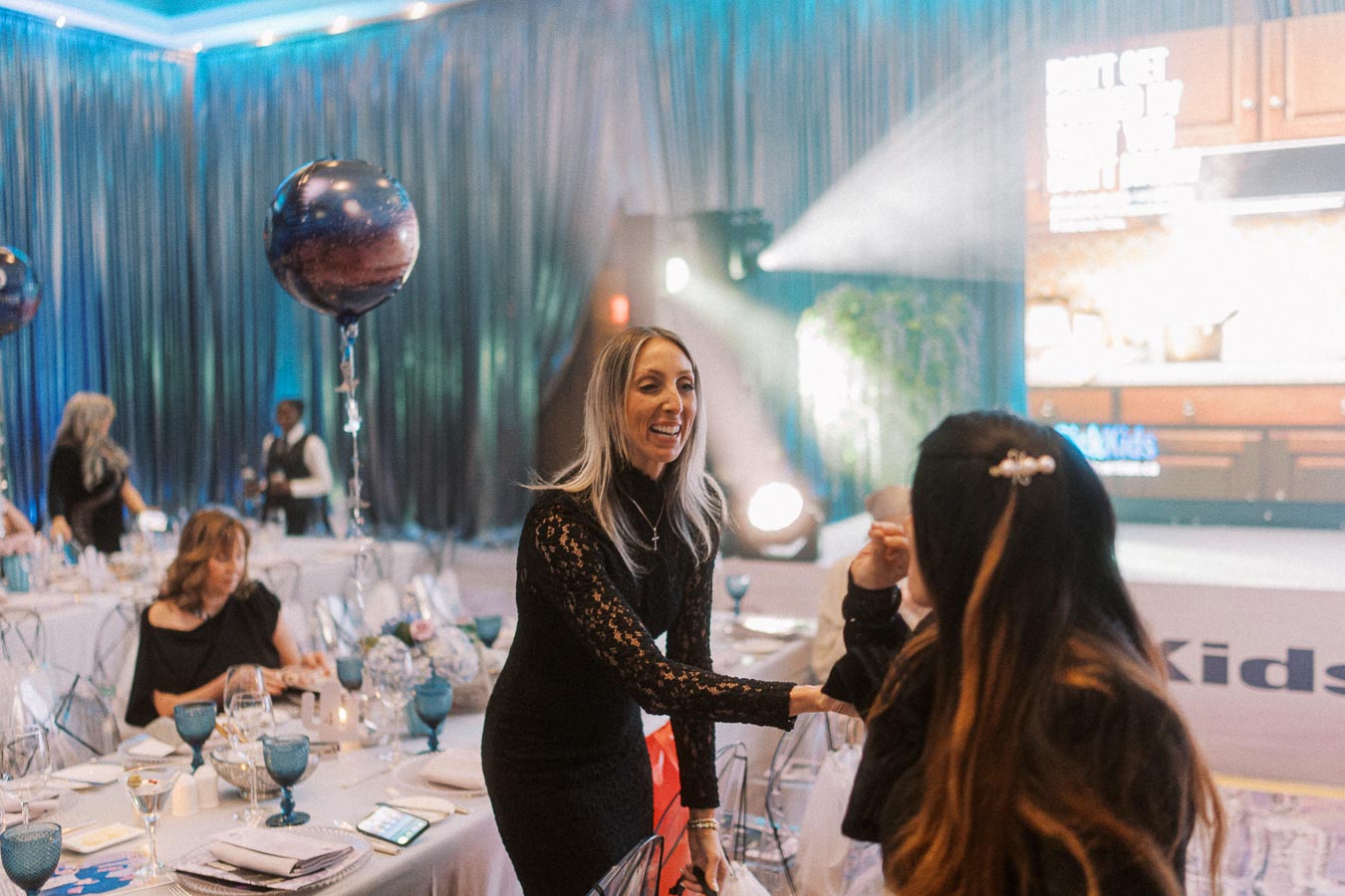 A woman in a black lace dress greeting a guest at an elegant event, with tables set for dining, blue goblets, and decorative balloons. A large screen is visible in the background, creating a sophisticated and inviting atmosphere.