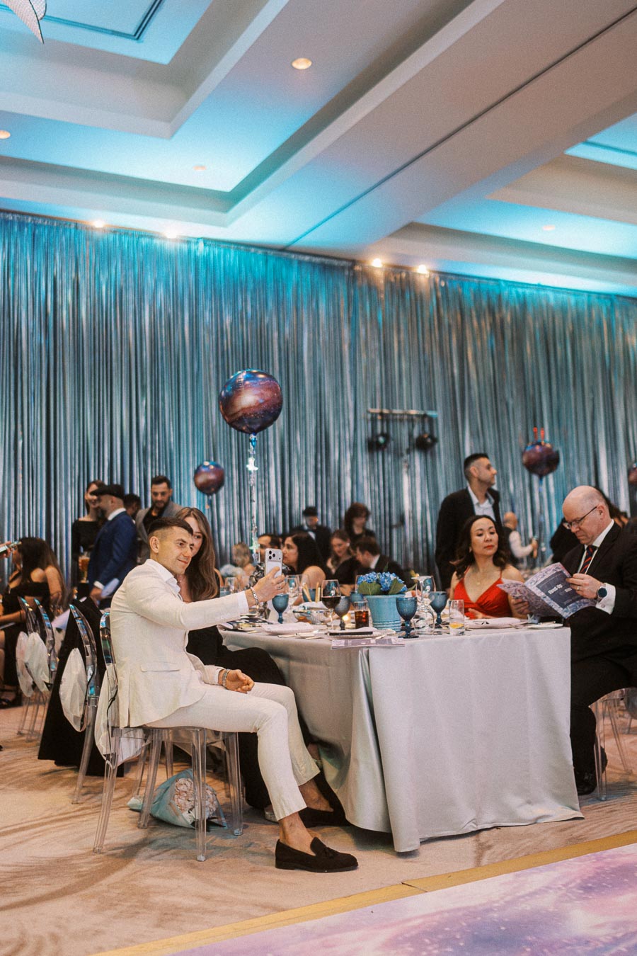 Guests enjoying an elegant event, with a man taking a selfie at a table adorned with aqua tableware and galactic-themed balloons under ambient blue lighting.