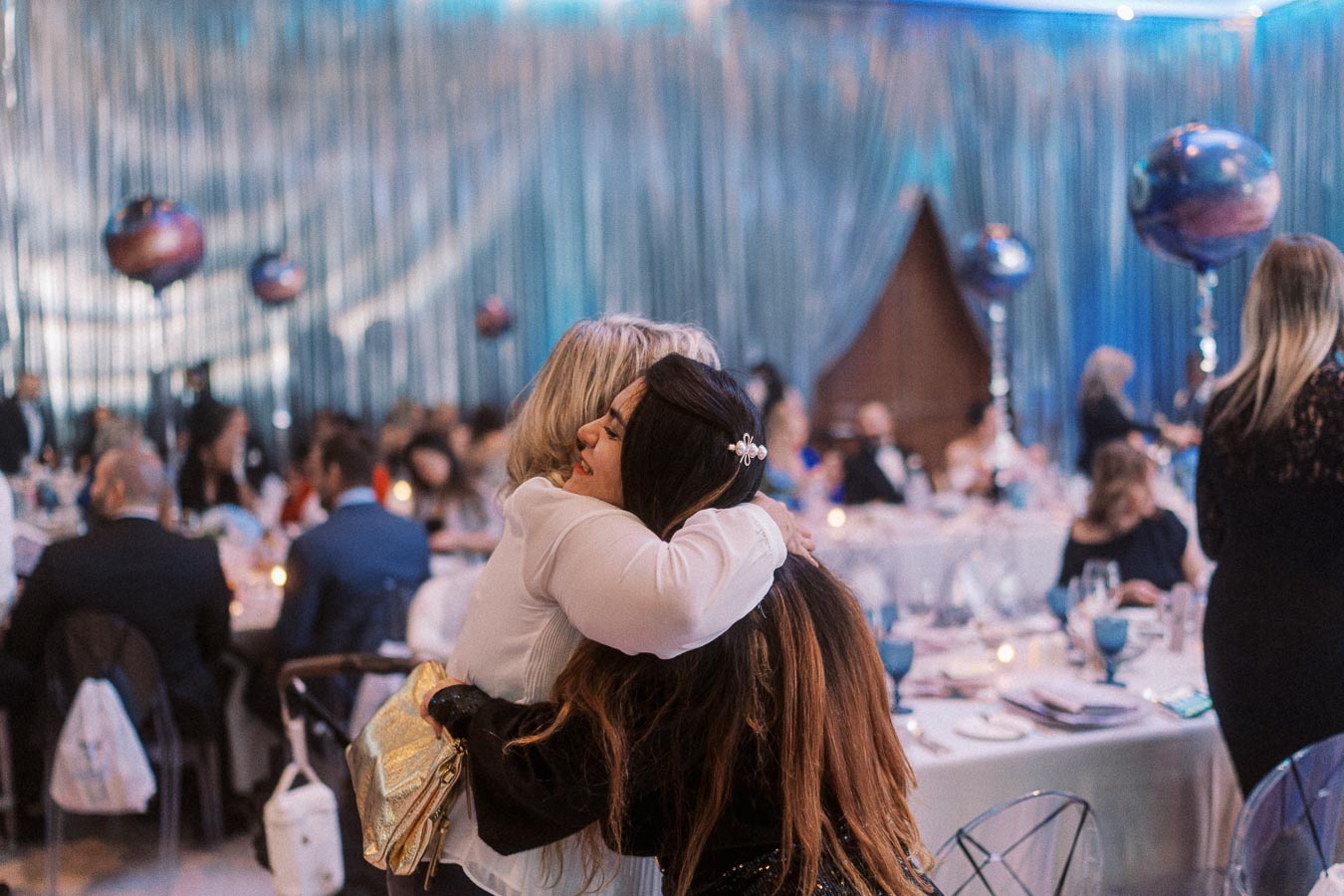 Two women embracing at an elegant indoor event with decorated tables and blue drapery, creating a warm and celebratory atmosphere.