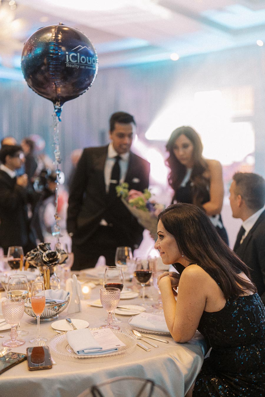 A group of elegantly dressed individuals at a formal event, gathered around a beautifully set dining table, with a distinctive balloon featuring iCloud Realty branding floating nearby.